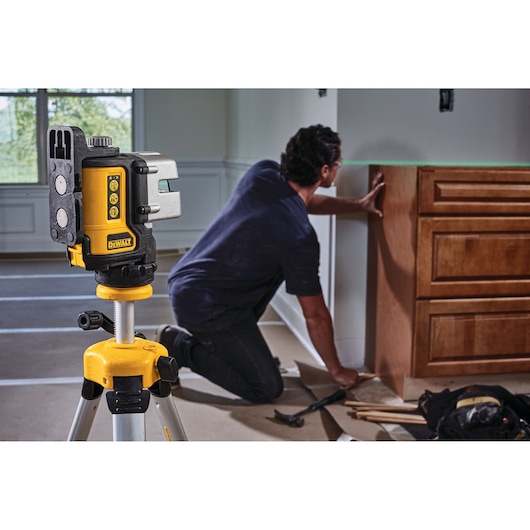 A DEWALT green laser level (model DW089CG) is mounted on a yellow tripod in the foreground, with a person kneeling in the background installing wooden cabinetry in a room under construction.