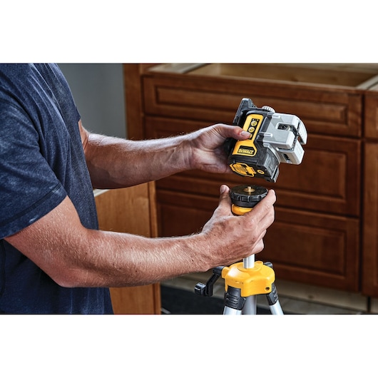 A person adjusts a DEWALT DW089CG green laser level mounted on a tripod, with wooden cabinets in the background.