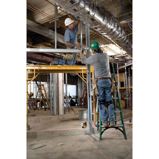 Two construction workers are installing metal framing in an indoor building site. One worker stands on a ladder, while the other is kneeling on a scaffold. Both are wearing safety helmets and tool belts. A DEWALT laser level (DW089K) is placed on the floor, projecting visible red lines onto the concrete for precise alignment.