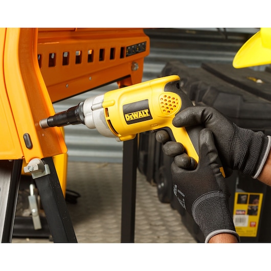A person wearing black gloves uses a yellow DEWALT electric screwdriver to fasten a bolt on an orange metal frame. The scene is set in a workshop with tools and equipment visible in the background.