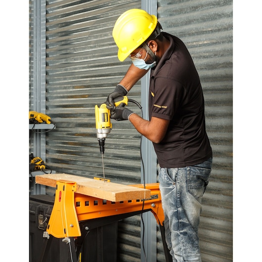 A person wearing a yellow safety helmet and gloves is using a yellow DEWALT electric drill to bore a hole in a wooden plank that is secured on a workbench inside an industrial workshop.