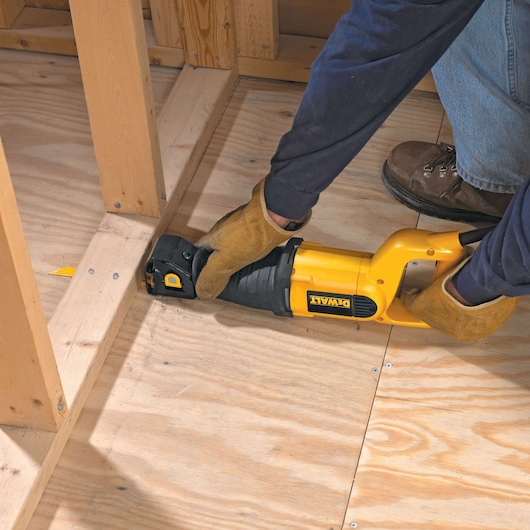 A person wearing gloves and work boots is using a yellow DEWALT reciprocating saw to cut wooden boards on a plywood floor during construction.