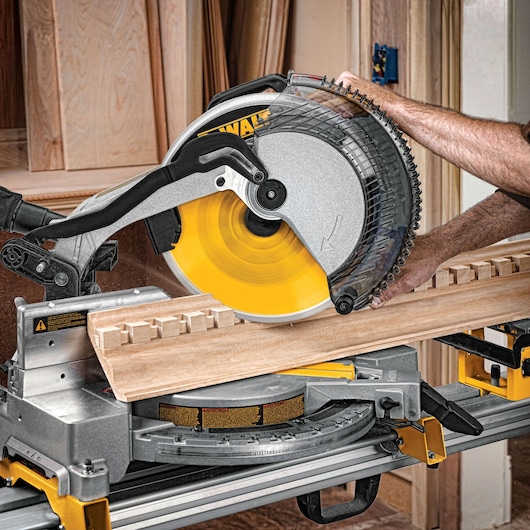 A DEWALT miter saw cutting a piece of wood, operated by a person, with wooden panels in the background.