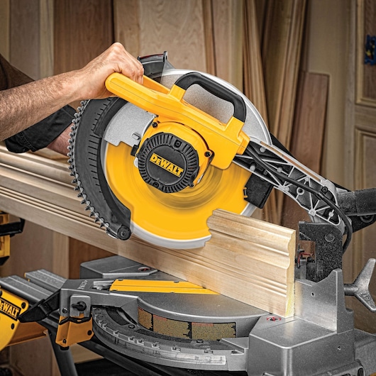 A DEWALT miter saw cutting a piece of wooden molding in a workshop. The saw is being operated by a person and the brand name is clearly visible on the tool.
