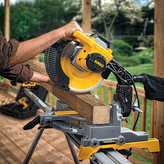 A person is using a DEWALT miter saw to cut a wooden beam on a deck outdoors. The saw is mounted on a stand and the DEWALT brand logo is visible on the saw. The setting includes natural greenery in the background.