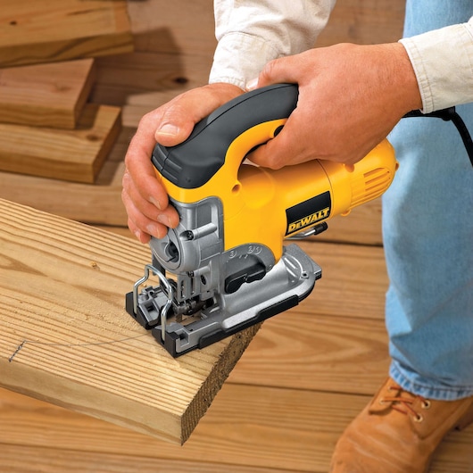 A DEWALT DW331K jigsaw being used to cut a wooden board, held by a person wearing a long-sleeve shirt and work boots. Several wooden planks are visible in the background.