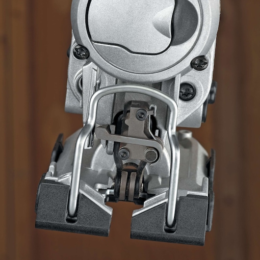 Close-up view of the lower part of a DEWALT jigsaw power tool, showing its metal blade guard, adjustment mechanism, and base plate against a blurred brown background.