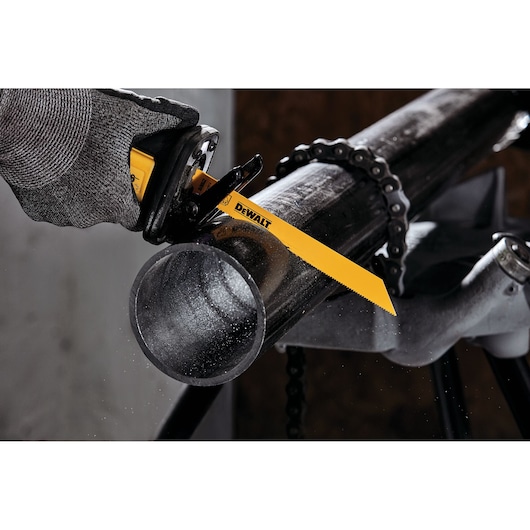 A close-up of a person using a DEWALT reciprocating saw blade to cut through a metal pipe. The person is wearing a protective glove and the pipe is secured in place with a clamp chain on a metal stand.