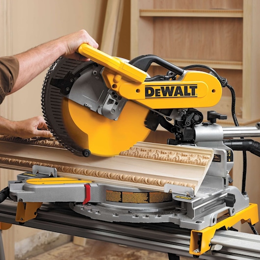 A Dewalt DW7084 miter saw being used to cut decorative wooden trim in a workshop setting. The saw is yellow and silver, and a person's hand is guiding the wood under the blade.
