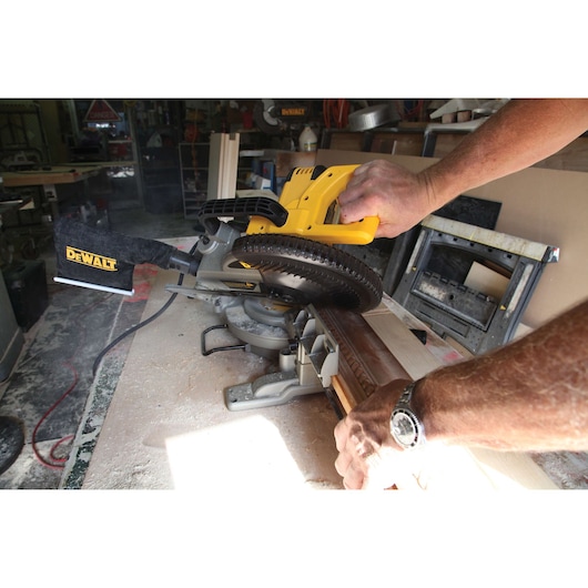 A person is using a DEWALT DW714 miter saw to cut a piece of wood in a workshop. The saw is yellow and black, and there is sawdust on the workbench. The person's hands are holding the wood and operating the saw.