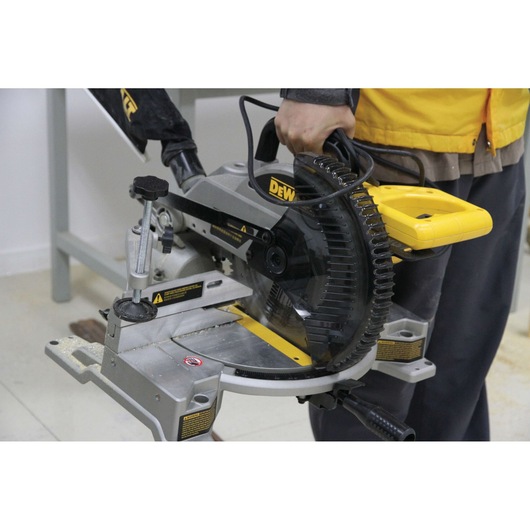 A person holding a DEWALT DW714 miter saw in a workshop setting. The saw is positioned on a workbench, displaying its blade, handle, and adjustment mechanisms.