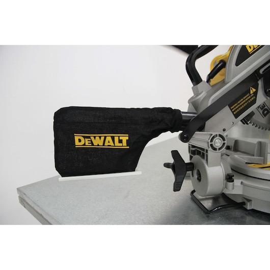 Close-up view of a DEWALT DW714 miter saw, focusing on the dust collection bag attached to the saw, placed on a metallic work surface.
