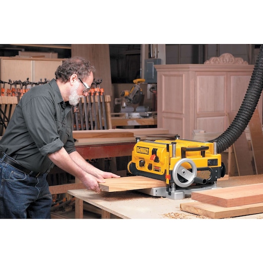 A person using a DEWALT DW735 thickness planer in a woodworking shop, feeding a wooden board through the machine. Various woodworking tools and materials are visible in the background.
