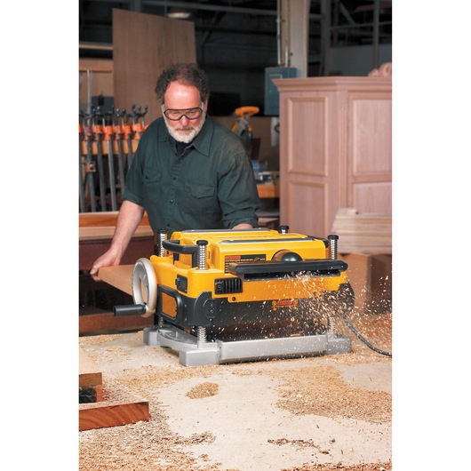 A person using a DEWALT DW735 planer in a woodworking workshop, with wood shavings being ejected from the machine. The person's face is blurred, and various woodworking tools and cabinets are visible in the background.