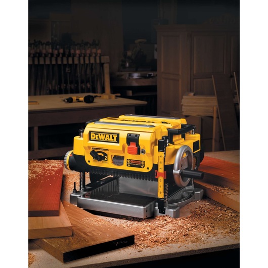 A DEWALT DW735 benchtop thickness planer is shown on a workbench in a woodworking shop, surrounded by wood shavings and wooden boards.