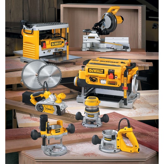 A collection of DEWALT woodworking power tools displayed on a workbench. The tools include a planer, a miter saw, a belt sander, circular saw blades, and several types of wood routers. The devices are yellow and black, surrounded by wooden boards and sawdust.