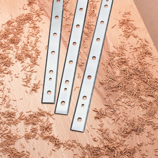 Three metal planer blades with several holes laid out on a wooden surface surrounded by wood shavings.