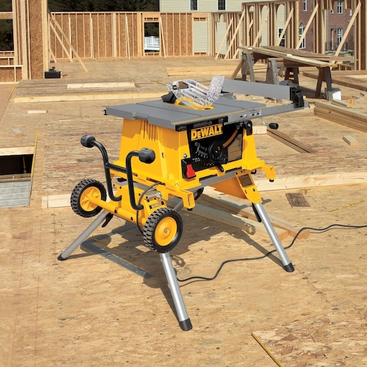 A DEWALT portable table saw with a stand and wheels is set up on a construction site with wooden framing and unfinished flooring.