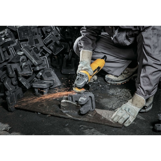 A person wearing protective gloves and work clothes is using a yellow DEWALT angle grinder to grind a piece of metal, producing sparks. There are several metal parts scattered on the floor around the person.