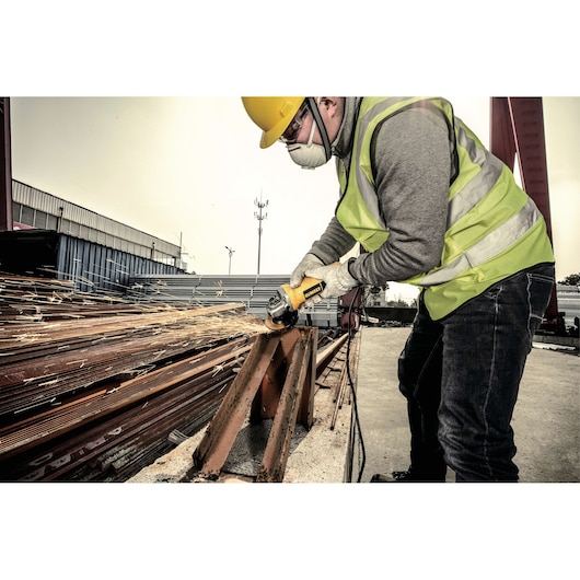 A worker wearing a safety vest, gloves, helmet, and face mask uses a DEWALT angle grinder (DW801) to cut a metal beam, producing sparks at an outdoor industrial site.
