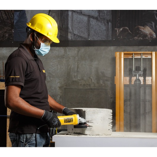 A person wearing safety gear, including a yellow helmet, black gloves, and a mask, operates a DEWALT DW802 angle grinder on a concrete block in an industrial or workshop setting.