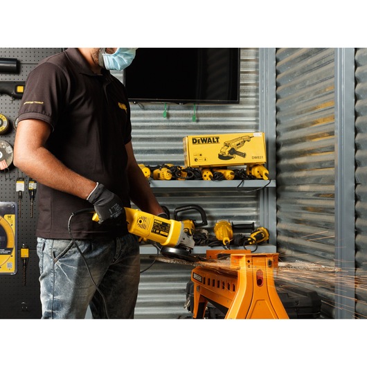 A person wearing a mask and gloves is using a yellow DEWALT angle grinder to cut metal on a workbench, with sparks flying. The workspace displays various power tools and a DEWALT DW831 product box on a shelf.
