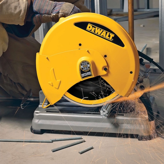 A person operates a DEWALT metal chop saw, model DW8500, cutting steel rods. Bright sparks are emitted as the rotating saw blade cuts through the metal. The tool features a yellow protective cover with the DEWALT logo.