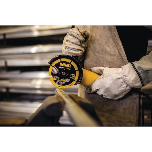 A person wearing protective gloves uses a DEWALT angle grinder with a yellow cutting disc to cut a metal pipe in an industrial setting.