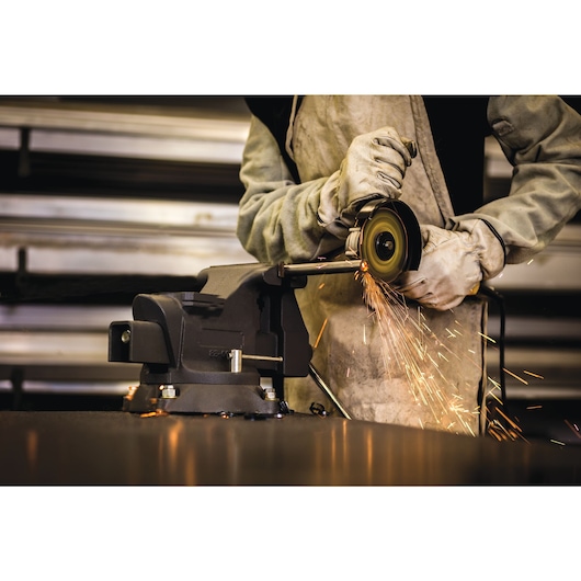 Person wearing protective gloves and work clothes using an angle grinder to cut metal, with sparks flying, in a workshop with a metal vise attached to a workbench.