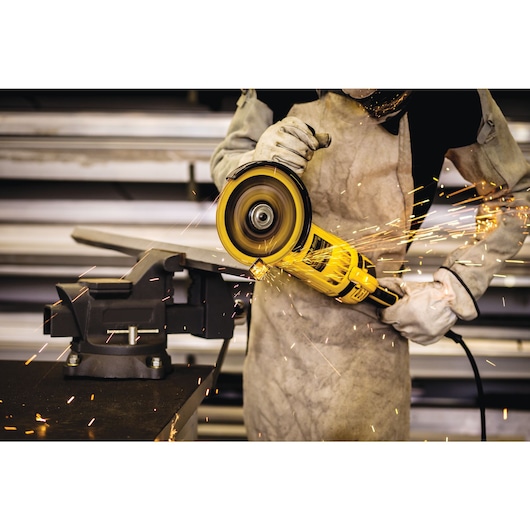 A person wearing protective gloves and clothing is using a yellow angle grinder to cut a piece of metal clamped in a vise, with sparks flying from the cutting process.
