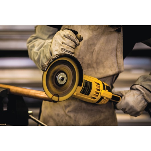 A person wearing protective gloves and an apron is using a yellow DEWALT angle grinder to cut through a metal pipe.
