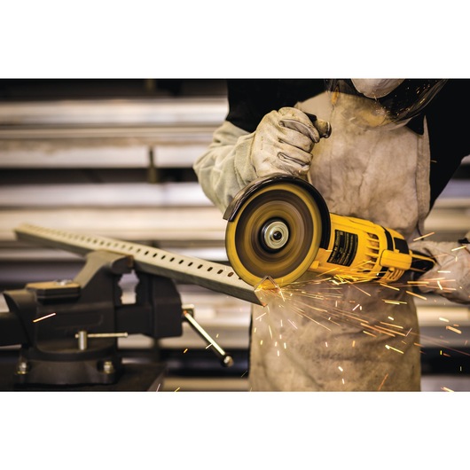 A person using a DEWALT angle grinder to cut a metal bar, with sparks flying. The person is wearing protective gloves, an apron, and a face shield.