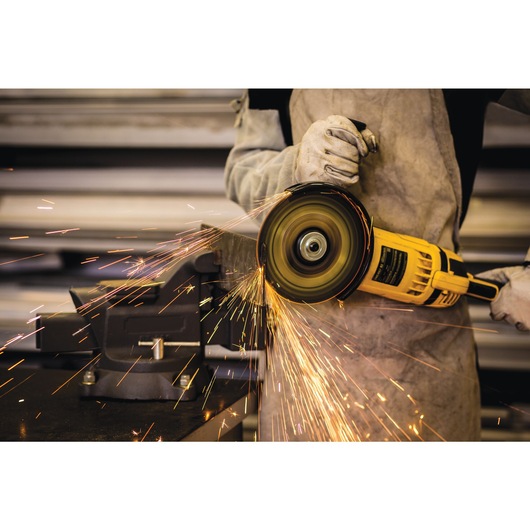 A person wearing protective gloves and an apron is using a yellow DEWALT angle grinder to cut a piece of metal clamped in a vice, creating bright sparks.