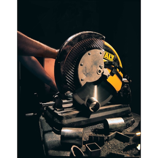 A DEWALT metal cutting saw is in use, cutting through a cylindrical metal pipe. The saw has a large circular blade and is mounted on a sturdy base. There are several pieces of cut metal and metal shavings scattered on the table around the saw.