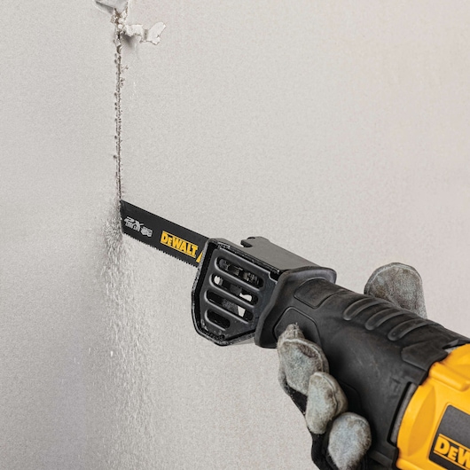 A close-up of a hand wearing work gloves using a DEWALT reciprocating saw to cut through drywall.