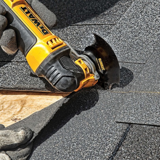 A close-up of a DEWALT oscillating multi-tool being used to cut roof shingles. The tool is held by a gloved hand, and the blade is making a precise cut on the shingles above wooden roof decking.