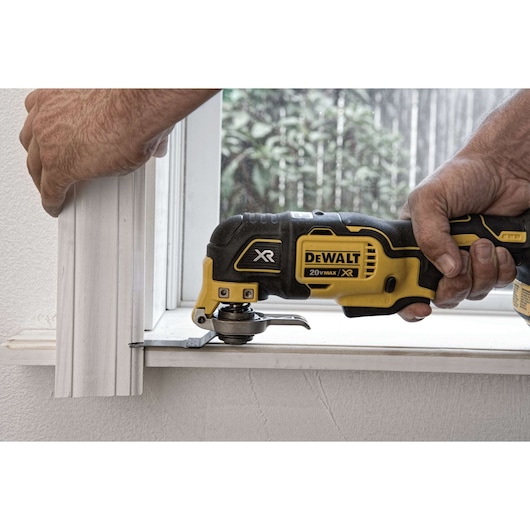 A person uses a DEWALT XR 20V Max oscillating multi-tool to cut a piece of window trim. The tool is yellow and black and being held close to a window.