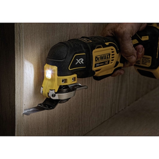 A DEWALT XR 20V Max oscillating multi-tool is being used to cut into a wooden surface. The tool's LED light is illuminated, and a person is holding the tool.