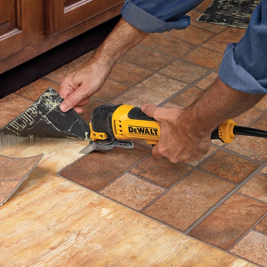 A person uses a yellow DEWALT oscillating multi-tool to remove a section of flooring tiles. The tool is cutting through the tile near a wooden cabinet, revealing the adhesive and bare floor underneath.