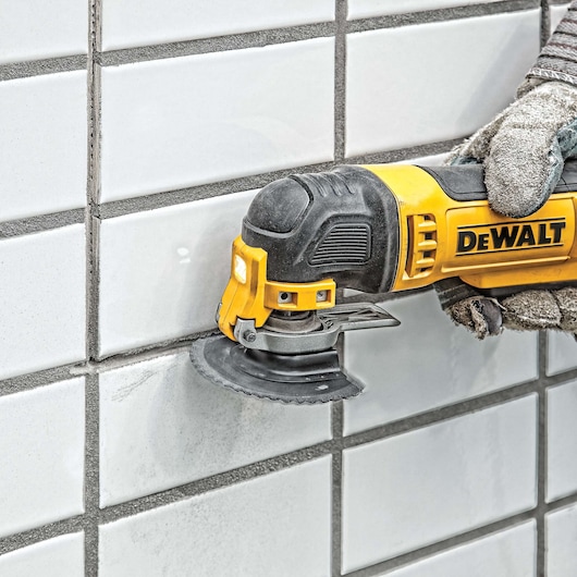 A gloved hand uses a DEWALT oscillating tool with a circular blade to cut or sand the grout lines between white ceramic tiles on a wall.