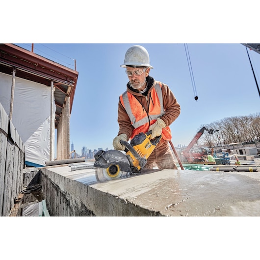 A construction worker wearing a hard hat and safety vest is using a DEWALT DWAFV8900 power saw to cut through a concrete slab at an outdoor construction site.