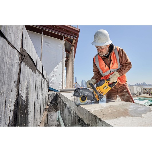 A construction worker wearing a white hard hat and orange safety vest uses a yellow DEWALT power tool to cut through a concrete block at an outdoor construction site. The worker's face is blurred for privacy. City buildings are visible in the background.