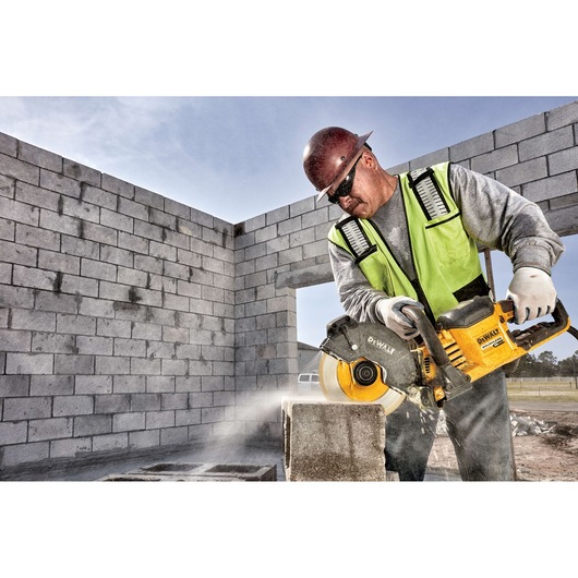 A construction worker wearing a safety vest and helmet uses a DEWALT power saw to cut concrete blocks on a building site. The image shows dust and debris flying while the worker operates the saw.