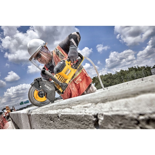 A construction worker wearing safety gear uses a DEWALT power saw to cut through a concrete block outdoors under a cloudy sky.
