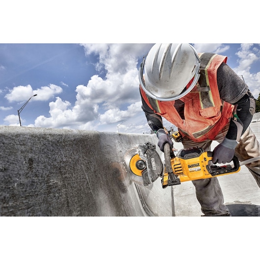 A construction worker wearing a safety helmet, gloves, and an orange safety vest is using a yellow DEWALT power tool to cut a concrete barrier outdoors under a partly cloudy sky.