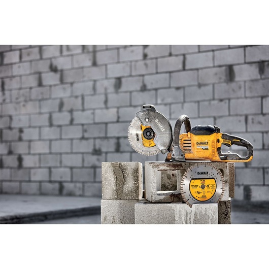 A DEWALT power saw and a circular saw blade are placed on concrete blocks in front of a gray brick wall.