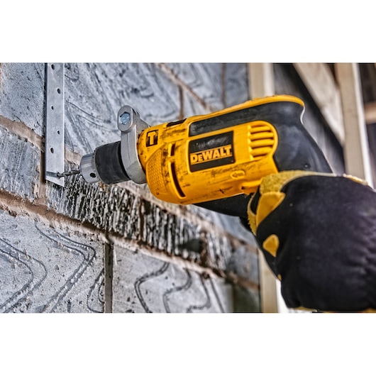 A person wearing work gloves is using a yellow and black DEWALT electric drill to drill into a brick wall near a metal bracket.