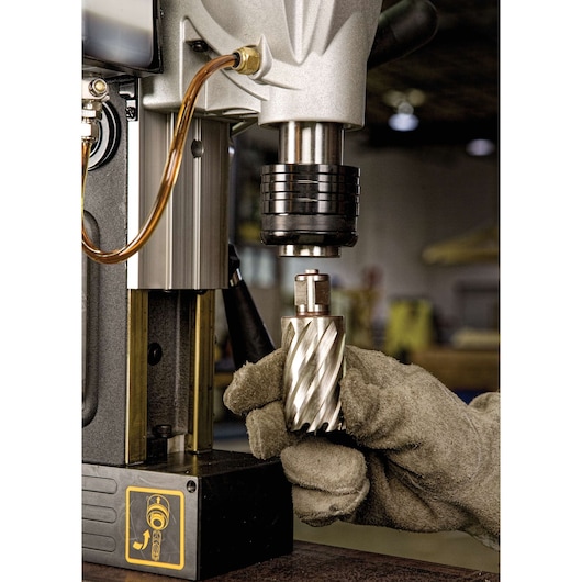 A close-up image of a worker's gloved hand inserting a metal drill bit into a DEWALT magnetic drill press. The scene is set in a workshop.