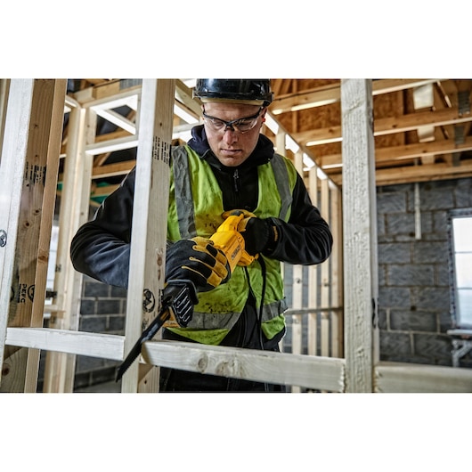 A person wearing a safety vest, gloves, and helmet uses a yellow DEWALT reciprocating saw to cut wooden framing at a construction site. The person's face is obscured.