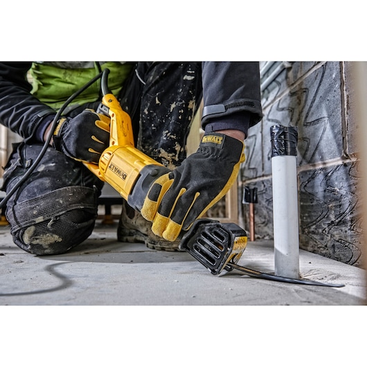 A person wearing work gloves and kneepads is using a DEWALT DWE305PK reciprocating saw to cut a metal pipe on a construction site floor.